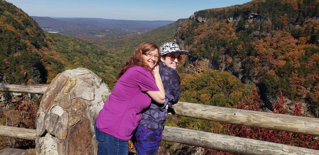Lorene and her daughter smiling together at a mountain overlook, surrounded by colorful fall foliage and forested cliffs.